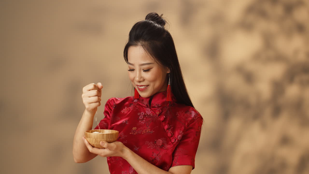 접시를 들고 손에서 쌀을 붓는 빨간색 전통 의상을 입은 쾌활한 젊은 아시아 여성