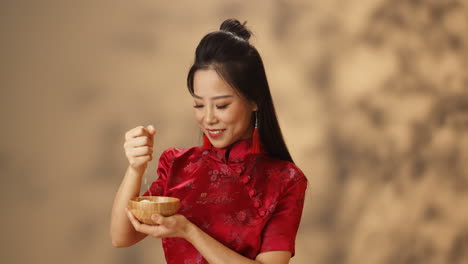 cheerful young asian woman in red traditional costume holding plate and pouring rice from hand