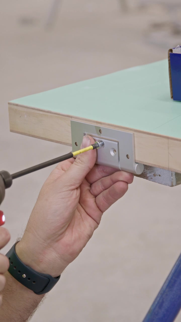 Primo piano verticale di una mano che usa un cacciavite per montare una cerniera metallica sul bordo di una porta di legno, tenendo ferma una vite durante l'installazione precisa di ferramenta per porte