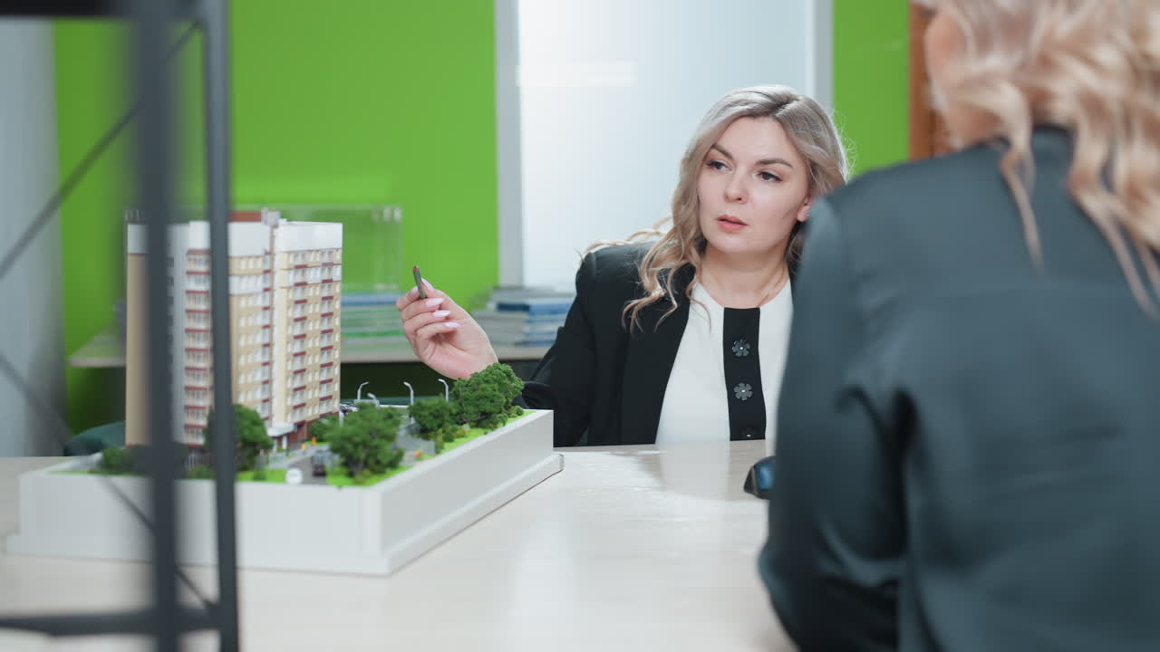 Realtor explaining architectural development project to client with professional demeanor, using pencil to highlight building model details during indoor meeting at desk with green background