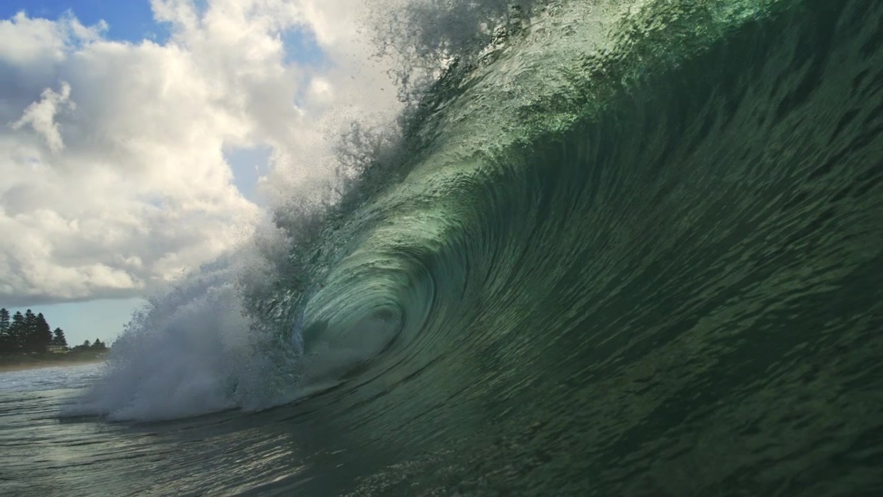 Green ocean wave rises over rippling water beneath sunlit clouds in steady slow motion