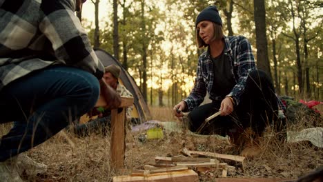 A-girl-in-a-black-hat-with-a-bob-hairstyle-helps-a-man-with-Black-skin-in-a-checkered-shirt-chop-wood-with-a-hunting-knife-during-a-camping-stop-in-a-sunny-summer-green-forest