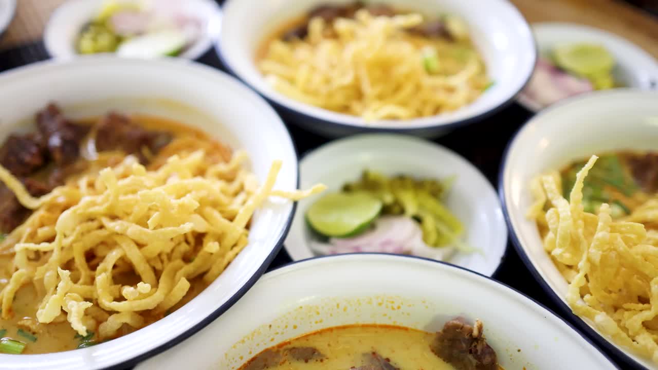 Multiple bowls of Northern Thai beef curry noodles with crispy toppings and side garnishes, shot in natural light with shallow depth of field