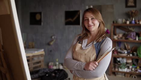 Portrait-shot-of-the-charming-and-pretty-caucasian-woman-artist-standing-crossed-arm-in-the-studio-among-pictures-with-brushes-in-hands-and-smiling-to-the-camera