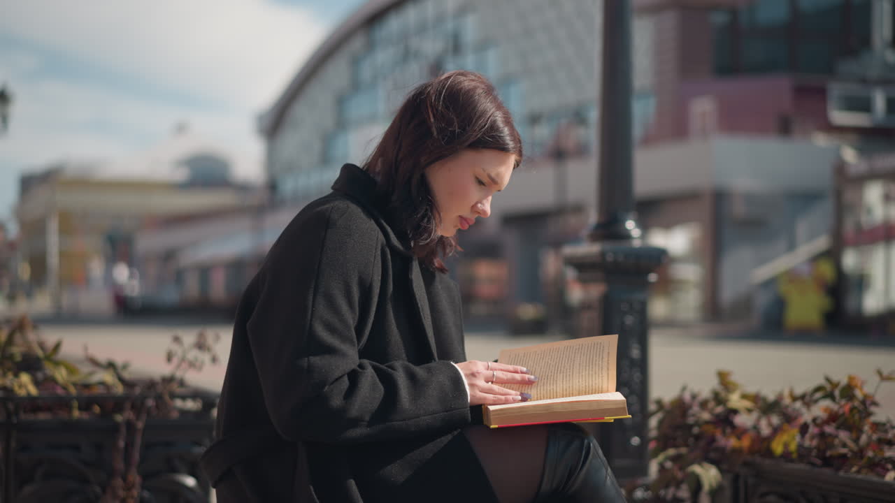 Young adult in black coat and leather boots sitting outdoors reading a book and flipping pages, with blurred street view and residential buildings in the background during bright sunny day