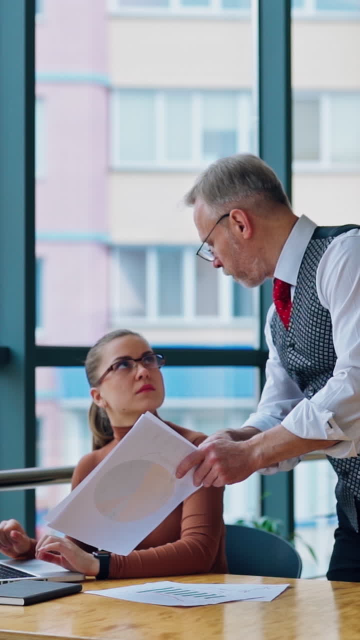 Senior businessman talking to the young secretary indoors. Serious woman in glasses listening to her old director near the window. Slow motion. Vertical video
