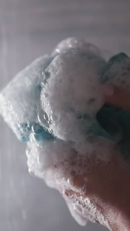 woman squeezes loofah in shower cabin closeup. lady hands hold foamy body washing sponge against water sprays. soft supply for personal hygiene