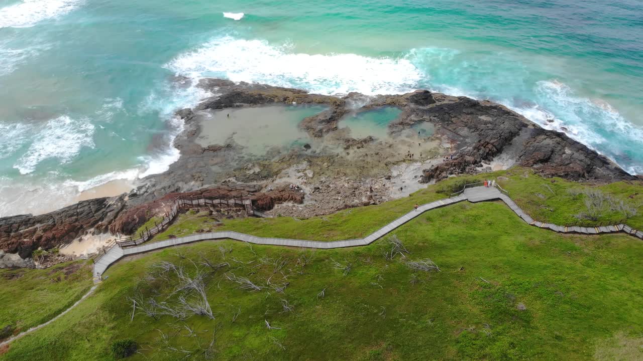 A drone captures a stunning view of tidal pools and waves crashing along a rugged coastline, with a winding boardwalk cutting through the vibrant green landscape at K'gari Island Champagne Rock Pools