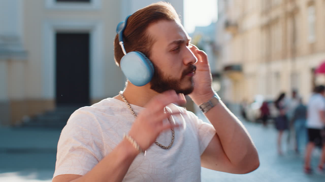 Happy young man guy in wireless headphones choosing listening music dancing outdoors city street