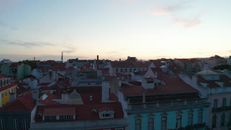 Drone-camera-starting-from-public-park,-rising-and-revealing-wide-town-panoramic-view-against-bright-sky-after-sunset.-Aerial-view-of-rooftops.-Lisbon,-capital-of-Portugal.