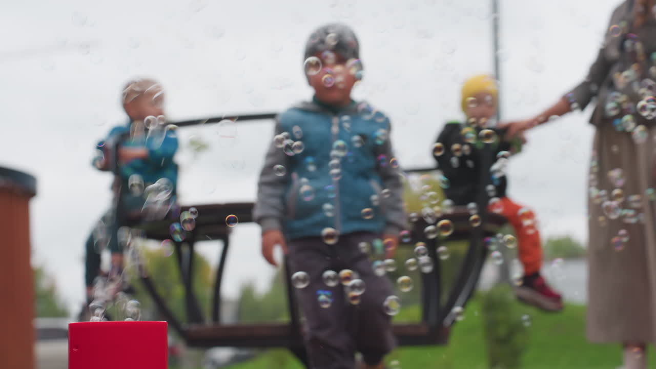 Soft focus park scene with child stepping off merry go round and walking toward red bubble machine, shimmering soap spheres drifting in air, caregiver nearby