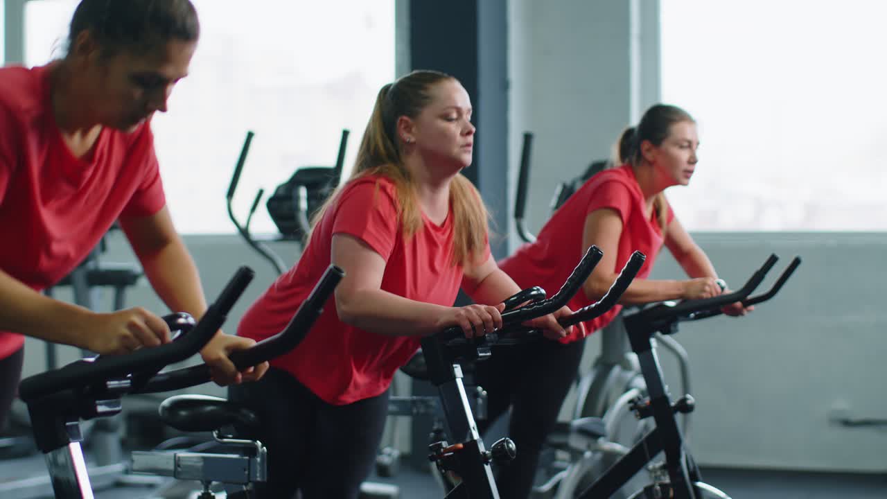 건강한 백인 여성 그룹이 체육관에서 정지 자전거 기계 자전거에서 운동을합니다.