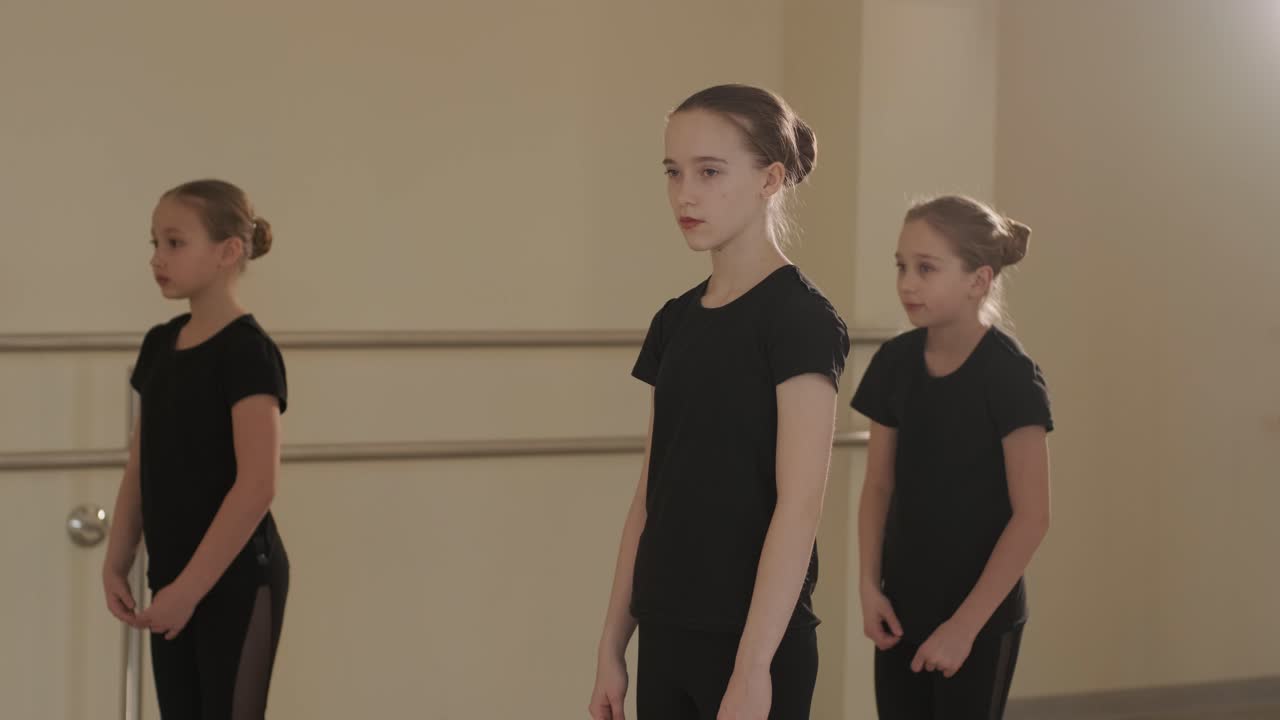группа молодых студентов балета в черной танцевальной одежде практикует позиции в просторной балетной студии с деревянным полом и настенными решетками. сосредоточенные выражения и синхронизированные движения.