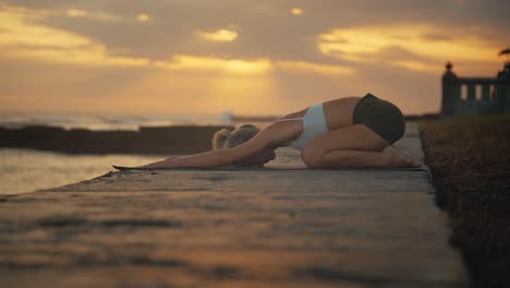 woman doing yoga child pose on shore of bali with sunset in background