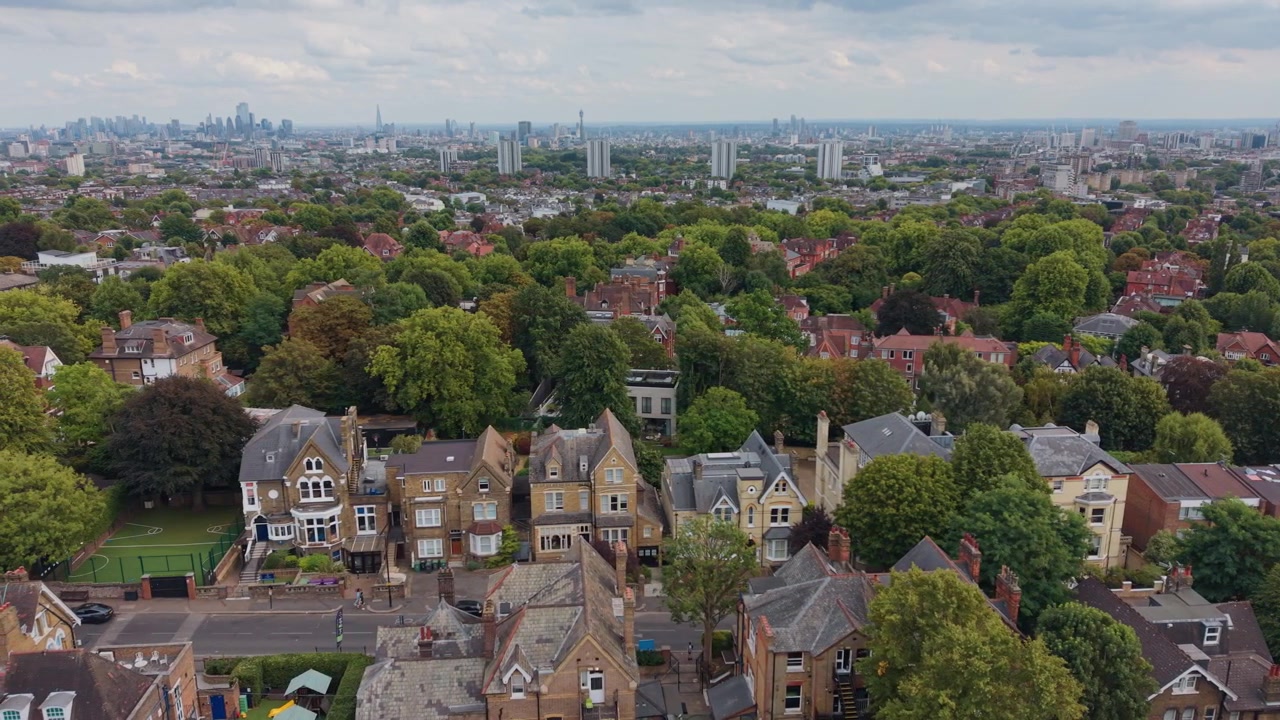 Leafy residential streets in North West London with large homes, apartment buildings, mature trees and the distant city skyline, aerial panning view