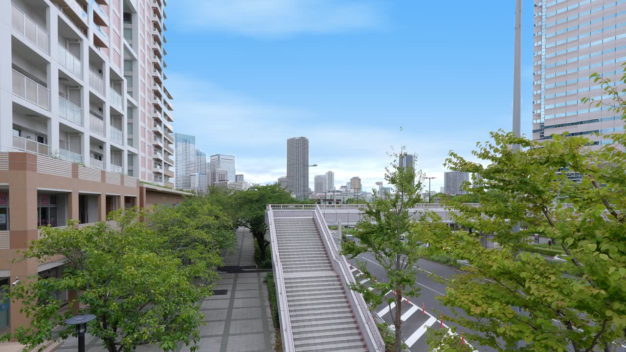 A wide shot of a modern urban park with a walkway and stairs, with the Tokyo skyline visible in the distance