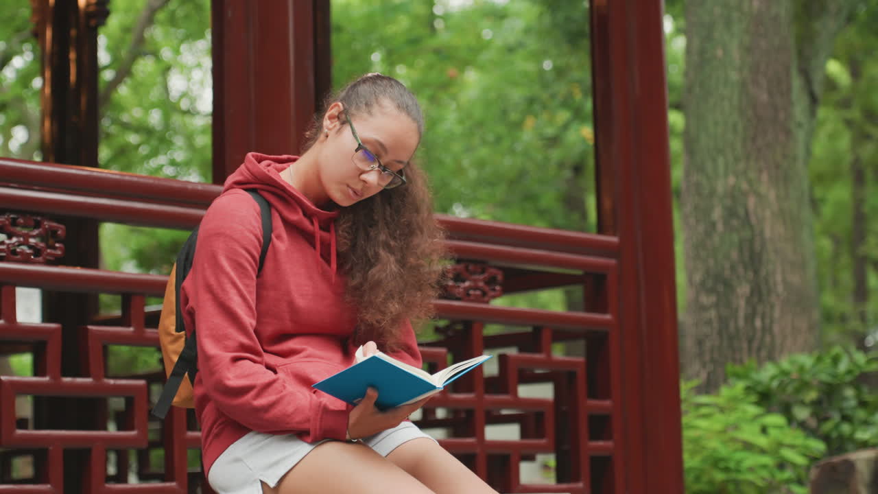 Weiße Frau studiert Notizbuch auf einer Bank, roter Hoodie und Rucksack neben der Schulter, Brille spiegelt Seiten wider, konzentrierter Ausdruck, Stift schwebt über Notizen, Atmosphäre der Prüfungsvorbereitung, hohe Bäume und Park