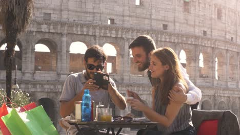 happy young couple tourists taking selfies with nice italian elegant waiter sitting at bar restaurant in front of colosseum in rome at sunset joking and laughing having fun