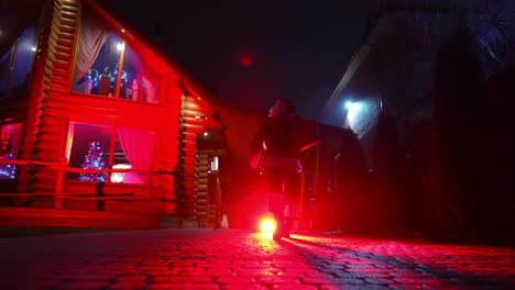 santa claus with bag full of presents. santa clause walking to the decorated houses with lights and carrying christmas gifts. red lighting. back view.