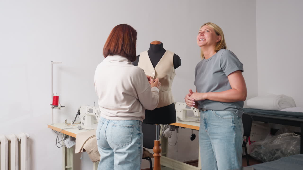 Garment makers converse cheerfully inside sewing studio as one buttons beige vest on black mannequin while other smiles, discussing fit details among industrial machines and folded fabrics