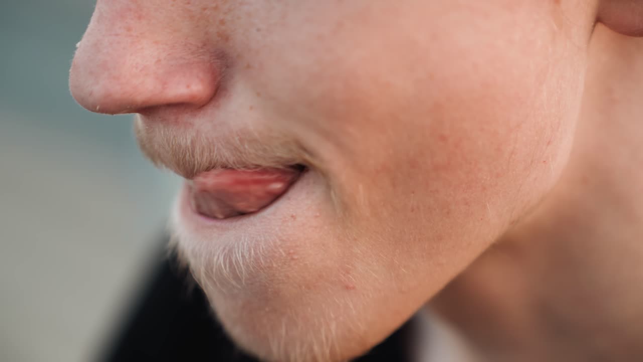 Primo piano di profilo laterale di uomo bianco che si morde il labbro, leggera spinta della lingua e sorriso nervoso, lentiggini e leggera barba incolta, sfondo sfocato del molo, luce serale, attore che ripassa le battute, palpabile anticipazione.