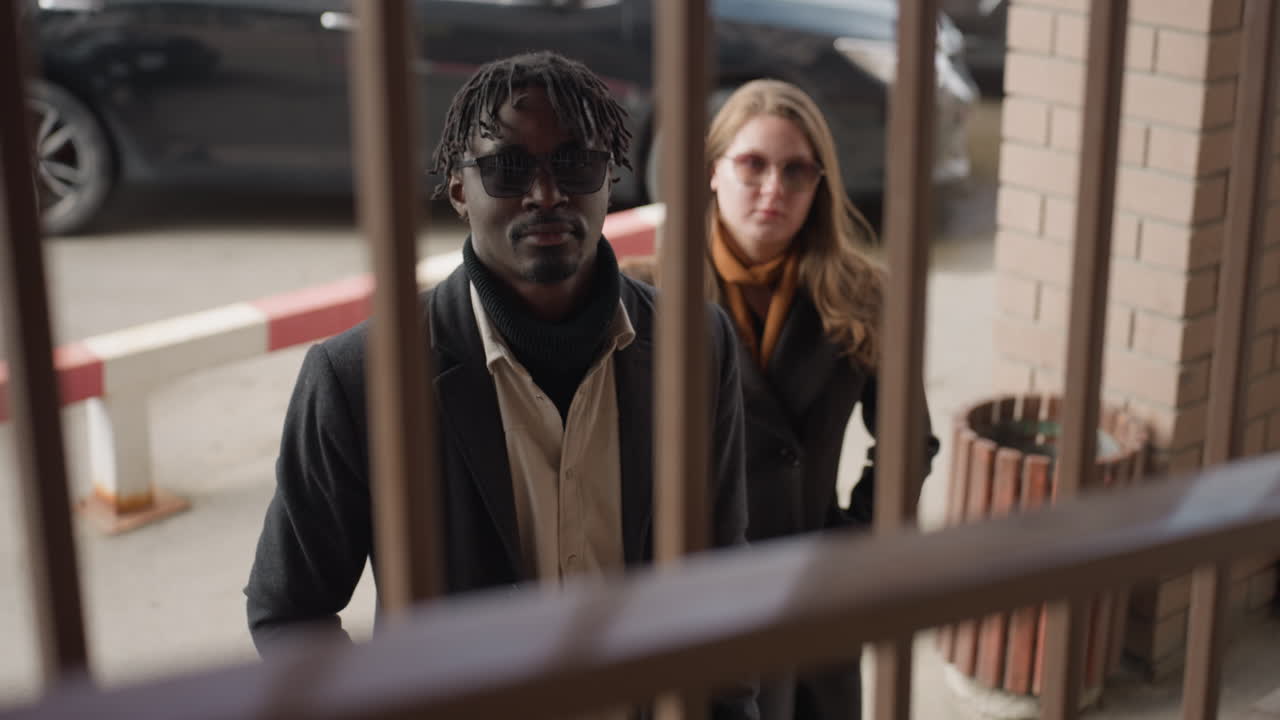 Black gentleman lowers head behind blurry iron barrier while friend stands softly focused in background, Scene shows subdued mood, muted daylight with parking barrier, candid moment framed by rail