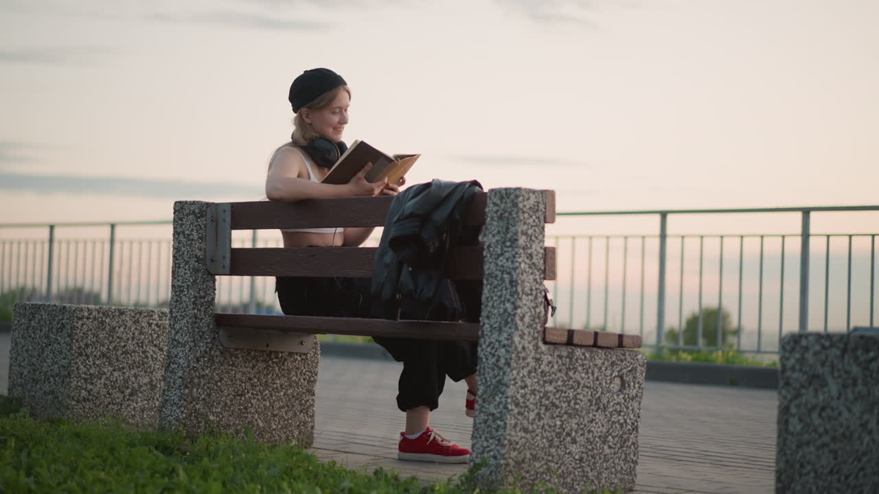 dançarino negro lendo em um banco à beira-mar ao pôr do sol, jaqueta jogada sobre o assento, tênis vermelhos visíveis, luz dourada suave, grade e horizonte distante, clima tranquilo e contemplativo após o ensaio, folheando