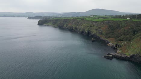 Aerial-view-over-the-cliffs-of-the-gorgeous-Kibane-Castle-in-Northern-ireland-along-the-giant-Causeway-coastal-road-overlooking-the-calm-blue-sea,-beautiful-scenery-and-mountains-in-the-background