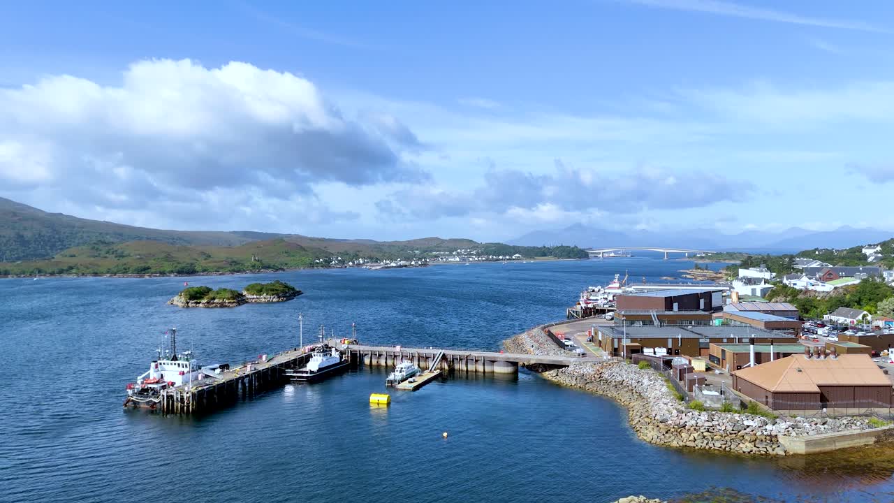 Aerial drone glides toward coastal village pier, revealing waterfront buildings, calm sea, and hills