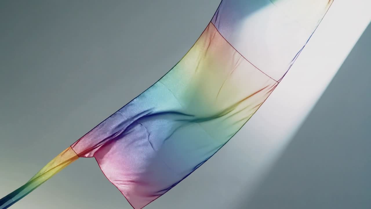 Unfurling long sheer rainbow fabric panel curling in room as diagonal beam rising, showing seams