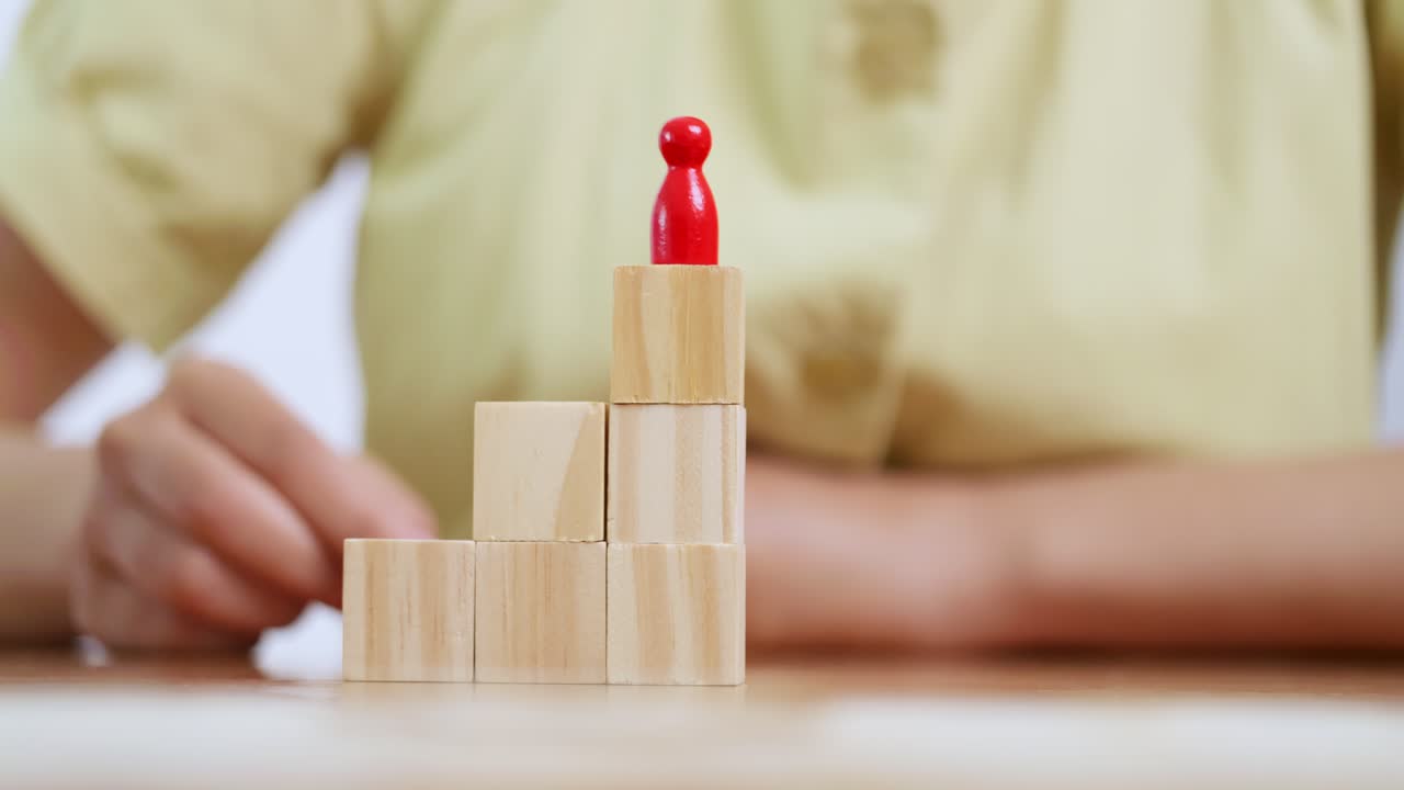 A person carefully places a red figure atop wooden blocks symbolizing growth and achievement