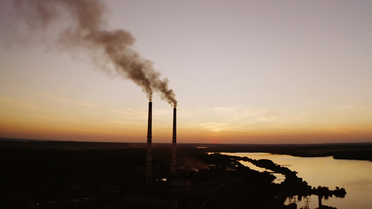 Manufacturing near the river at darkness. Two pipes with dirty smoke going into the air on the nature background in the evening.
