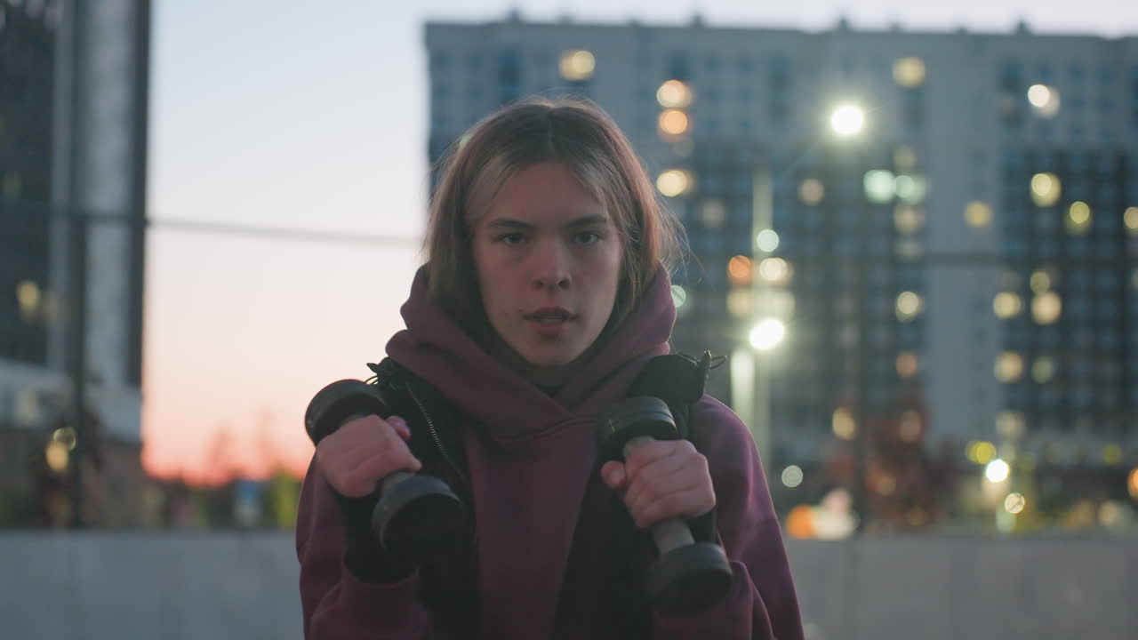 Portrait of gym lover with focused face boxing dumbbells punching air at dusk on black asphalt sports court next to white barrier and fence in urban setting wearing maroon hoodie and white sneakers