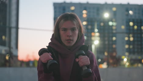portrait of gym lover with focused face boxing dumbbells punching air at dusk on black asphalt sports court next to white barrier and fence in urban setting wearing maroon hoodie and white sneakers