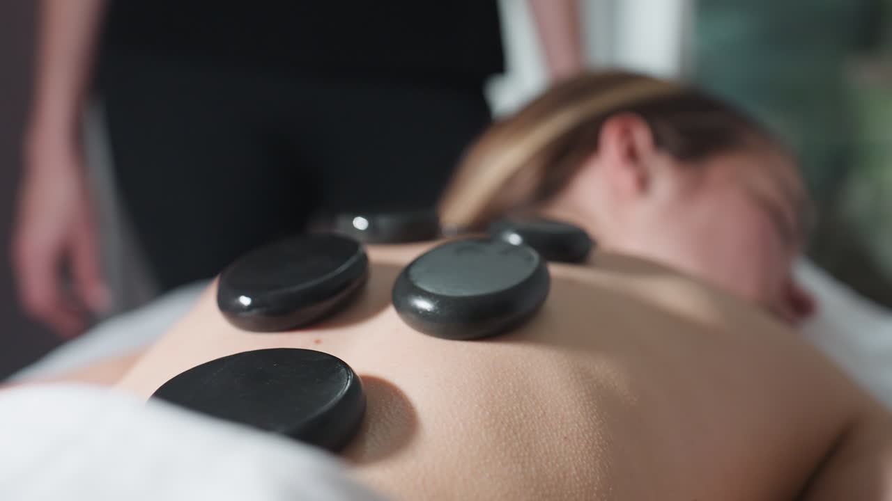 Very close up view of smooth massage stones placed on client back during relaxing therapy session with blurred background of massage therapist standing nearby, capturing warmth and healing ambiance