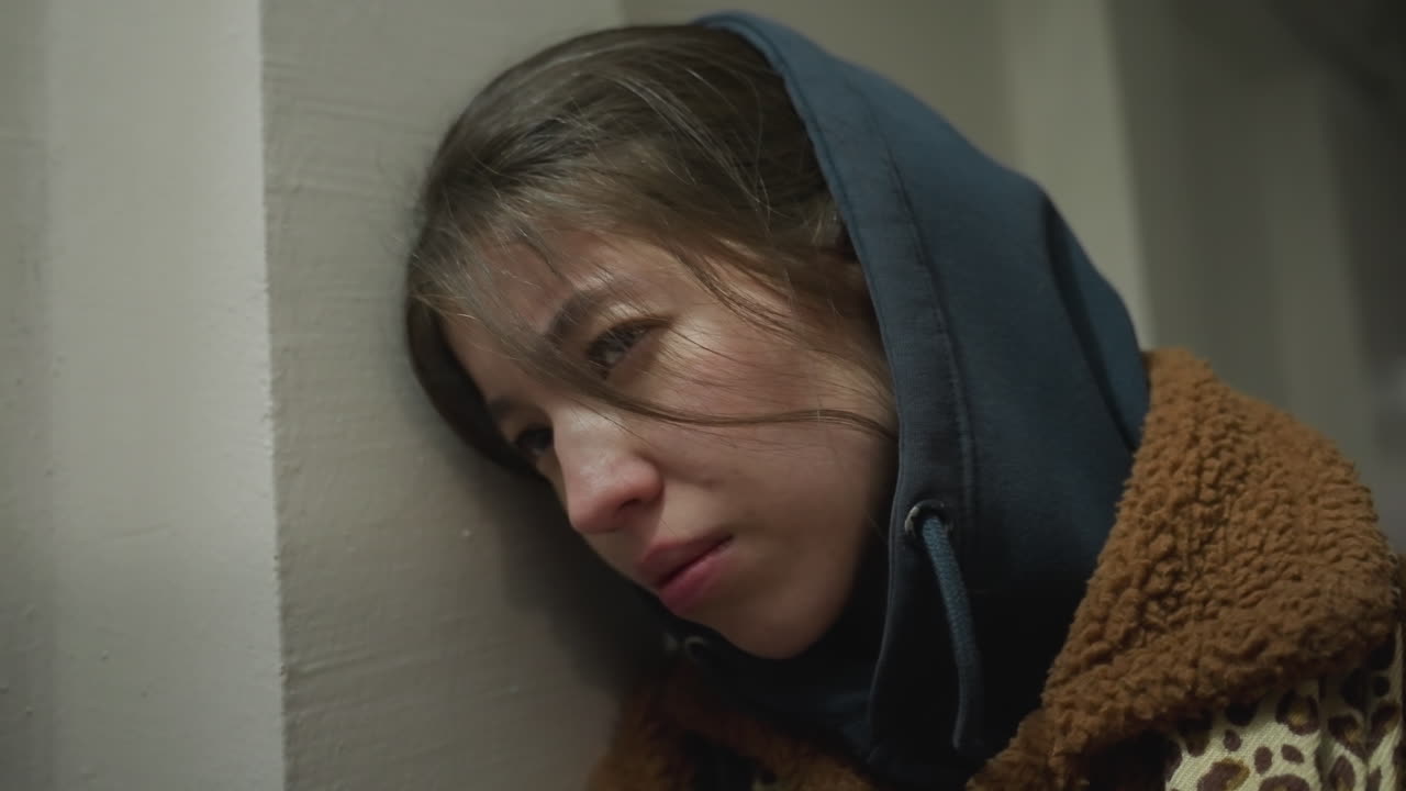 Close-up shot of a distressed girl in a brown coat, leaning against the wall of an underpass tunnel. She appears troubled, with a tense expression and her hoodie up, a mood of emotional turmoil