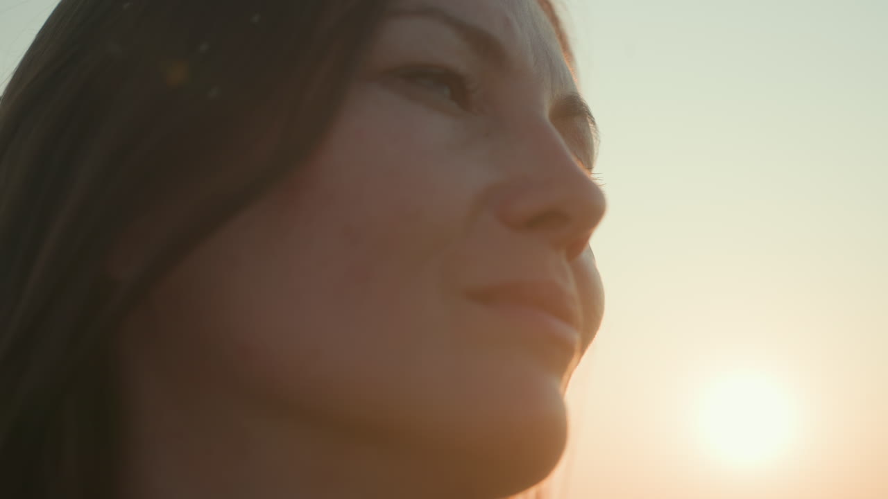 Close up of young woman staring into distance during sunset, soft golden sunlight casting gentle glow on her face, contemplative expression, breeze moving hair, flying insects visible