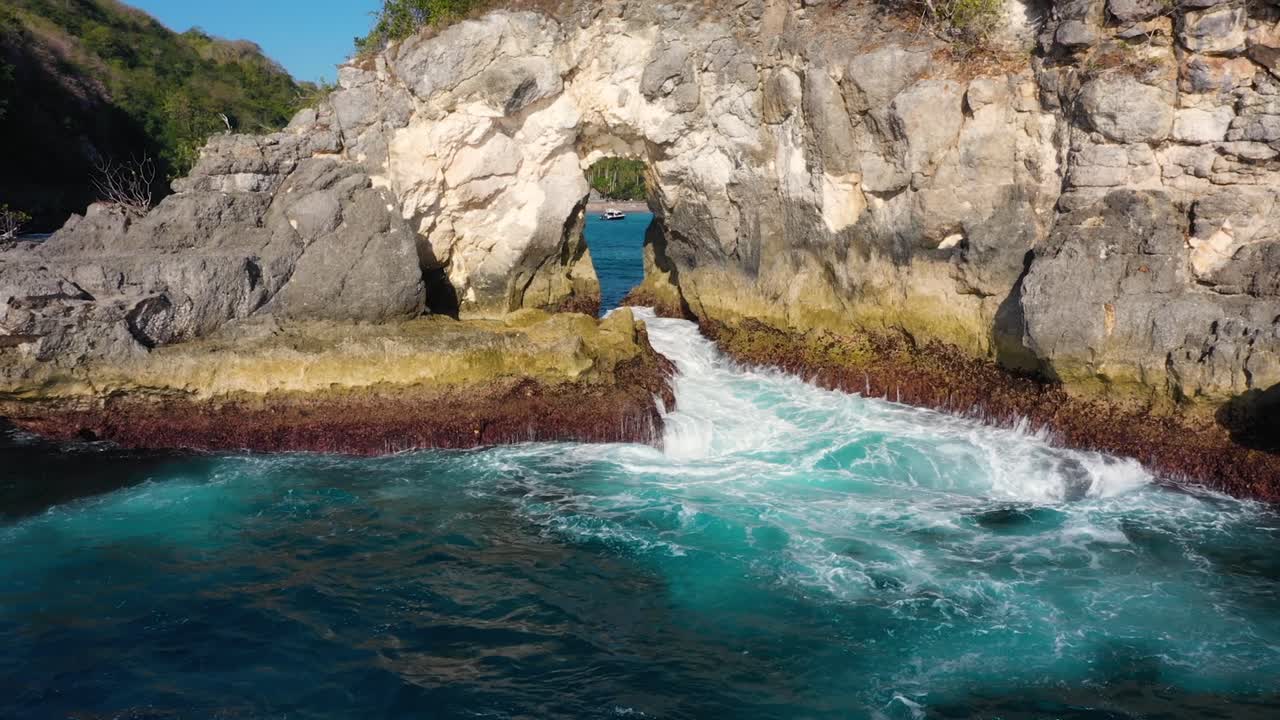 video aéreo cinematográfico de una isla tropical con olas de agua turquesa aplastando un arco rocoso con un bote en el fondo, playa de crystal bay, nusa penida, bali, indonesia