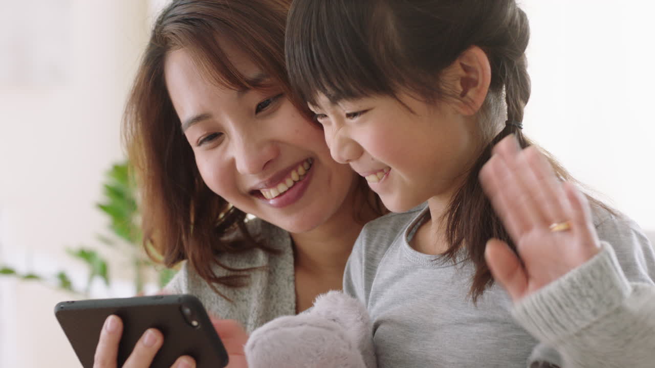 アジアの母親と子供がスマートフォンを使ってビデオチャットをしています 母親と小さな女の子が手を振って 娘と週末の休暇を共有しています 携帯電話でチャットを楽しんでいます 4k映像
