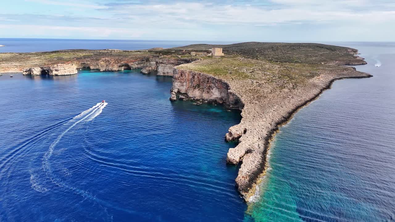 Flyfoto av robuste kystklipper og klart blått hav ved Lantern Point på Comino, Malta, som viser en smal steinete halvøy, små bukter og en ensom steinbygning.