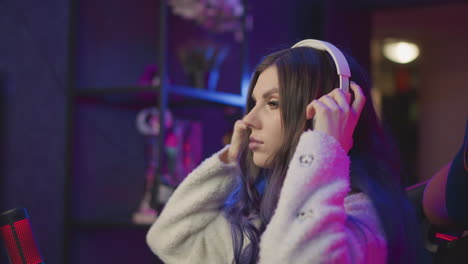 rear view of lady seated in gaming chair wearing fluffy jacket placing white headphone over ear while resting hand on desk near dual rgb monitors red microphone under vibrant neon lighting