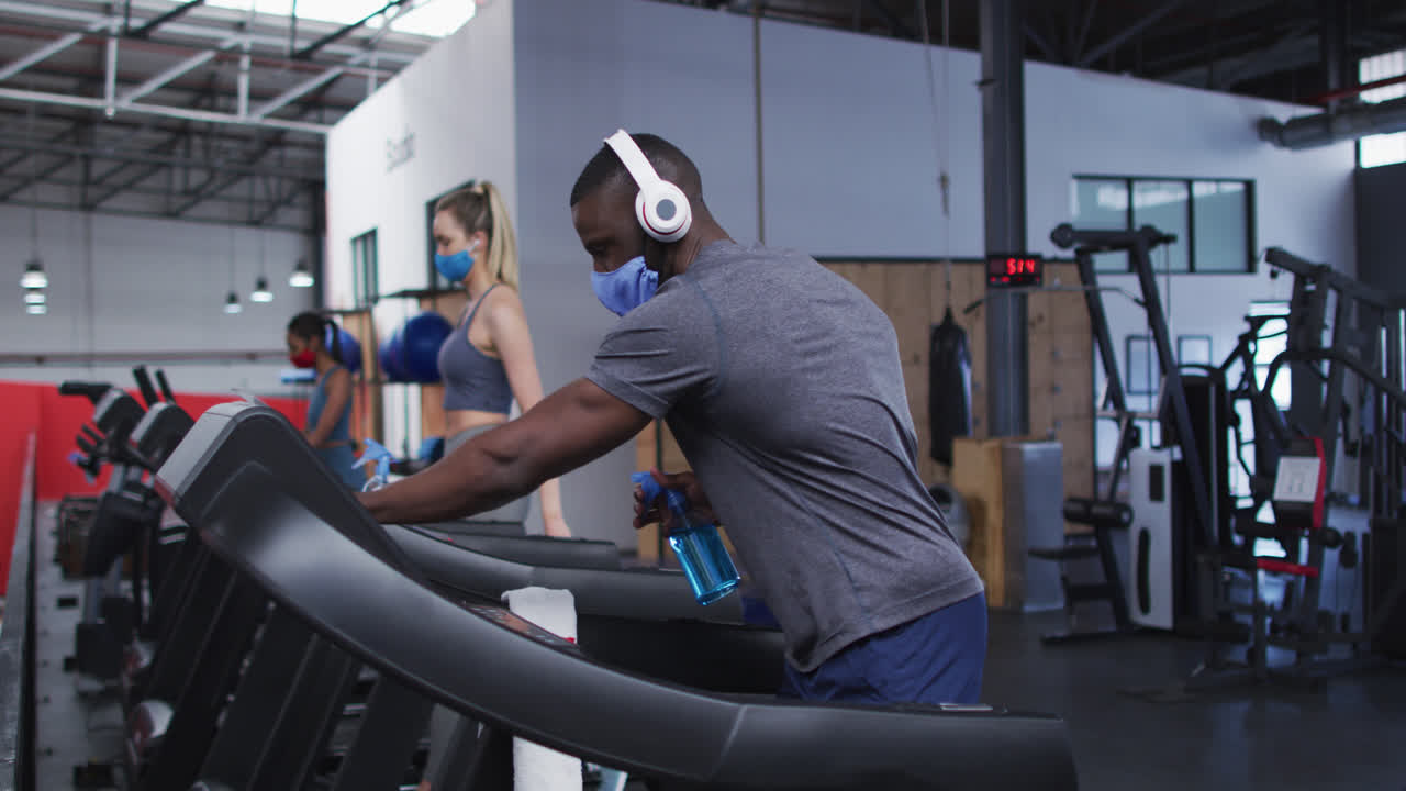 얼굴 마스크와 헤드폰을 착용한 건강한 아프리카계 미국인 남자가 소독제로 트레드밀 기계를 청소하고 있습니다.
