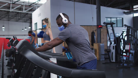 hombre afroamericano en forma usando máscara facial y auriculares limpiando la máquina de cinta con desinfectante