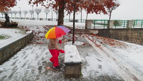 Niña-Pequeña-Con-Paraguas-De-Muchos-Colores-Juega-Pateando-Bolas-De-Nieve