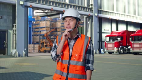 asian male engineer with safety helmet thinking about something and looking around while standing , outside of logistics distributions warehouse