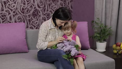 Little-daughter-and-mother-sitting-on-sofe-with-bouquet-of-lilac-purple-flowers