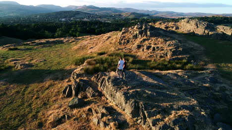 Drone-shot-circling-adult-couple-standing-on-a-rocky-peak-overlooking-Lake-Windermere,-Lake-District,-UK