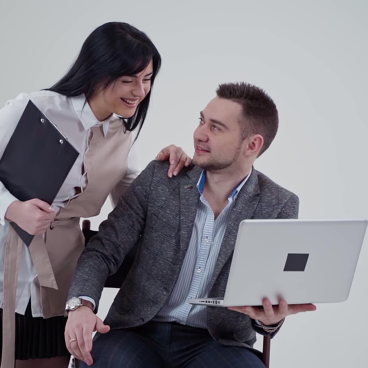 Beautiful couple in elegant clothing on white background. Handsome man sitting in chair and holding laptop. Young pretty woman standing close to a businessman.