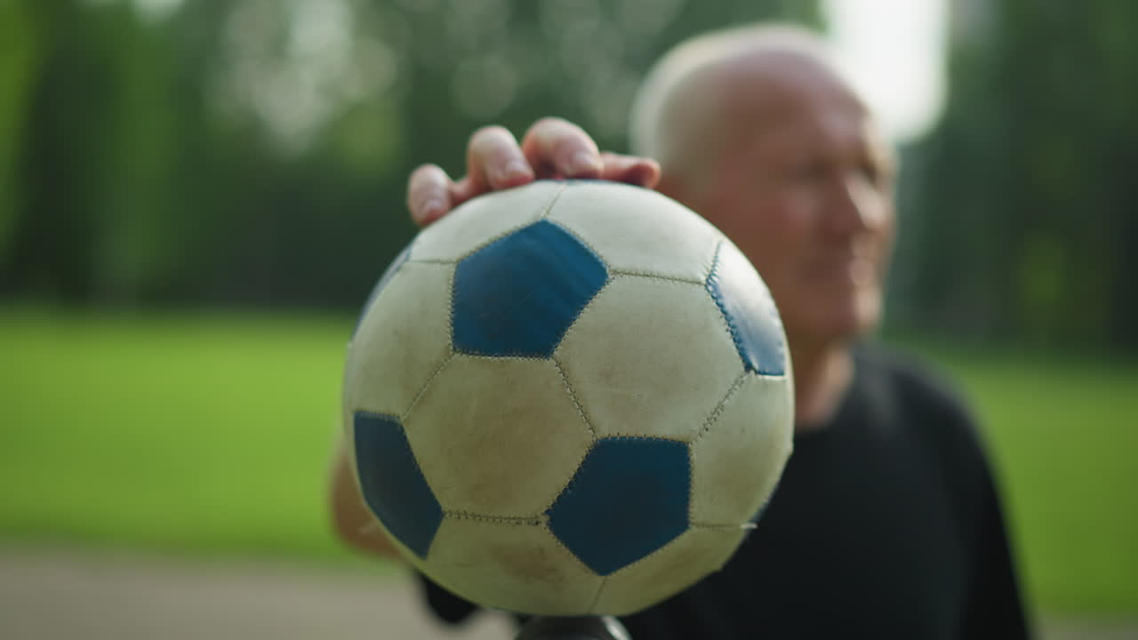 una vista borrosa de un anciano con una camiseta negra moviendo suavemente una pelota de fútbol con su mano, con una vista borrosa de una carretera pavimentada y un campo cubierto de hierba