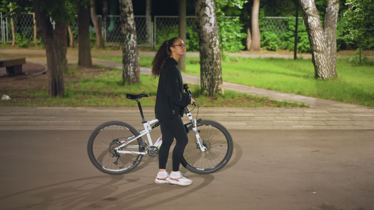 Gentle Evening Stroll Alongside Bicycle In Park, Woman Leisurely Biking Through Park During Quiet Evening Hours, Calm Nighttime Scene Of Woman Casually Cycling Past Playground Under Soft Lighting